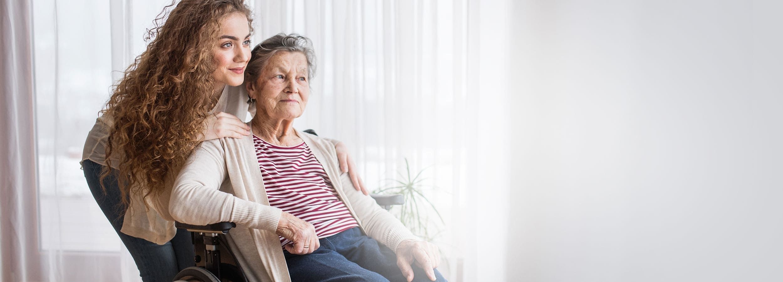 Junge Frau umarmt sitzende alte Frau von hinten